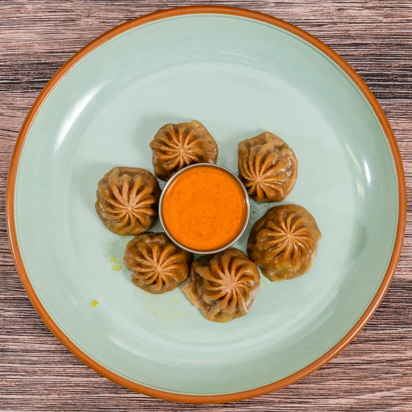 Six Indian momos with a vibrant orange dipping sauce in the center on a light green plate.