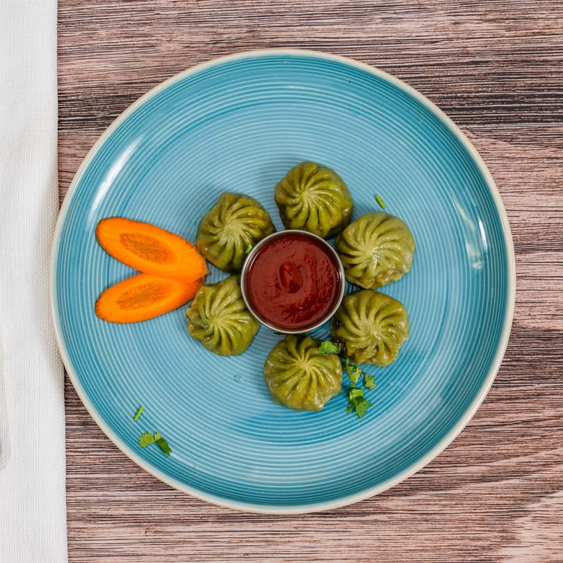 Green momos with red dipping sauce and carrot slices on a blue plate at Moh Moh Licious.
