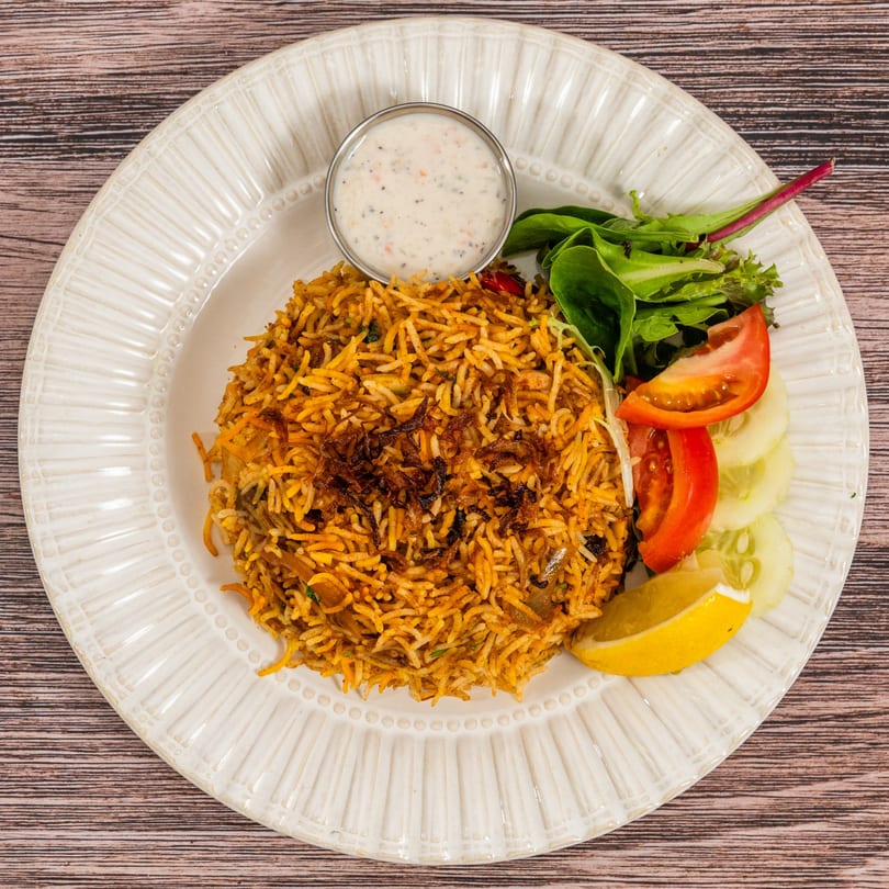 Indian biryani with fried onions, a side salad, cucumber, tomato, lemon, and raita.