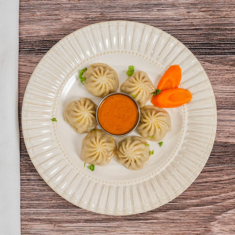 Six Moh Moh Licious momos with a side of orange dipping sauce and carrot slices on a white plate.